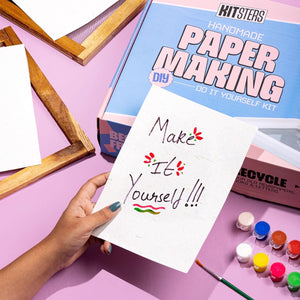 Handmade paper making kit with a hand holding a piece of paper with 'Make It Yourself' text, surrounded by paint bottles and a wooden frame on a pink background. Kitsters DIY Handmade Paper Kit at Go Science Product Image 8