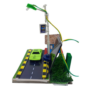 Solar Street Light School project model image 3 go science classroom series. A miniature diorama model featuring a toy green car on a road, a street light pole with wires, a small sign reading 'SAVE TWO LIFE', artificial grass, and a miniature building, all displayed on a white background.