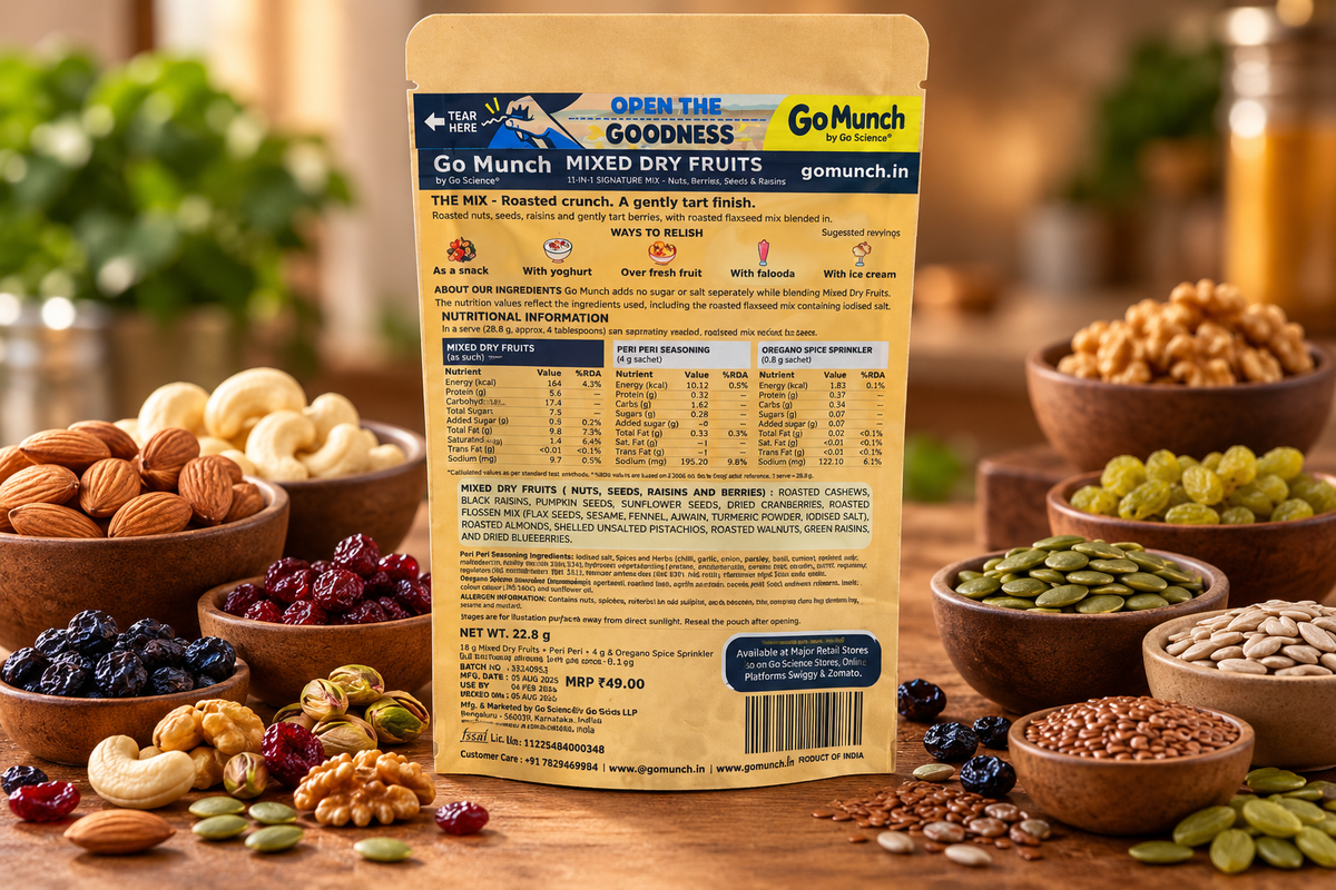 Back of a Go Munch Mixed Dry Fruits pouch shown on a wooden table surrounded by bowls of nuts, seeds, and dried fruits. The package is tan and blue, with nutritional facts and ingredients printed on the back.