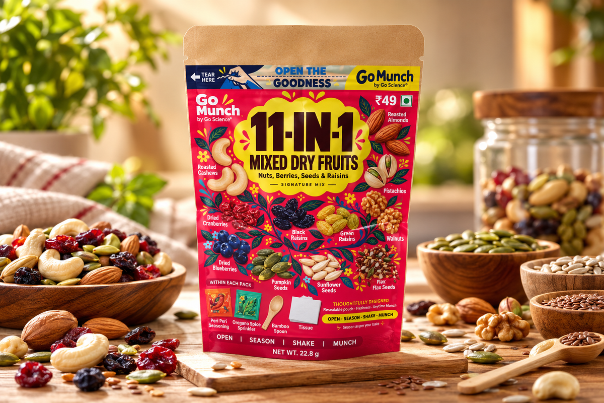 A stand-up pouch of Go Munch 11-In-1 Mixed Dry Fruits centered on a wooden table, surrounded by bowls of nuts, seeds, dried fruits, and jars in a warm kitchen-like setting. The package is bright red and yellow with multicolored fruit illustrations.