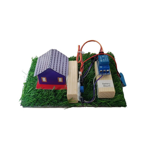 A small electronic model setup on a white background, featuring a miniature house with a purple body and gray roof, artificial green grass, two wooden blocks, wires, and a blue electronic component labeled 'Battery Mount'. From Go Science Classroom Series Fire Alarm Using Flame Sensor School Project & Demonstration Model kit 