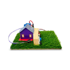A small toy house model with a purple body, blue-gray roof, and red base, mounted on a rectangular base with artificial green grass, a wooden plank, and visible electronic wiring on a white background.Go Science Classroom Series Fire Alarm Using Flame Sensor School Project & Demonstration Model kit  image 3