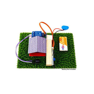 A DIY electronic circuit kit displayed on a square patch of artificial green grass, featuring a purple LED light block, a wooden block with a metal rod, a battery, wires, and a blue connector, on a white background with a text note 'Battery Not Included'. Go Science Classroom Series Fire Alarm Using Flame Sensor School Project & Demonstration Model kit  image 4
