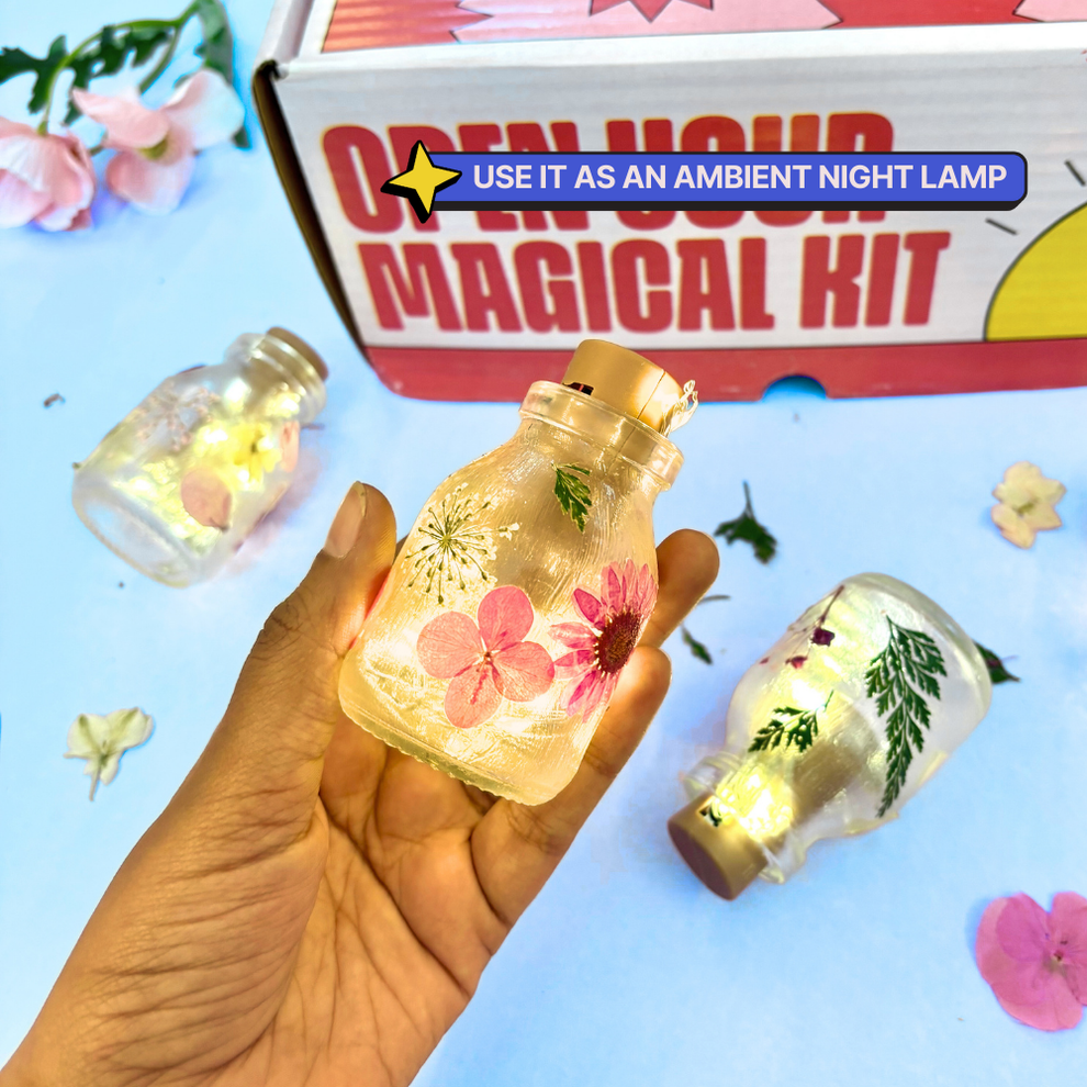 Hand holding a glowing bottle with floral designs, surrounded by more bottles and flowers on a light blue background. Kitsters DIY Floral Glow Jar Kit available at Go Science product image 3