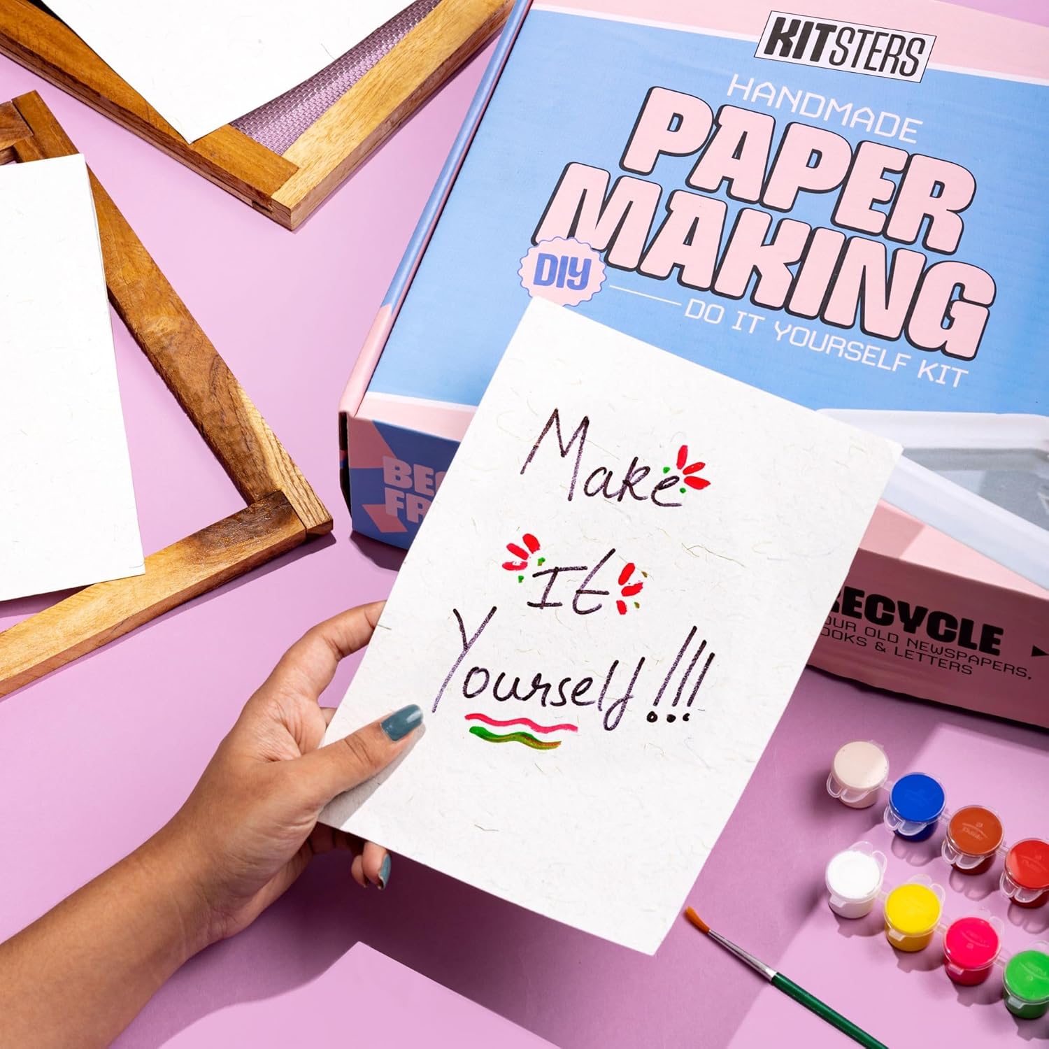 Handmade paper making kit with a hand holding a piece of paper with 'Make It Yourself' text, surrounded by paint bottles and a wooden frame on a pink background. Kitsters DIY Handmade Paper Kit at Go Science Product Image 8