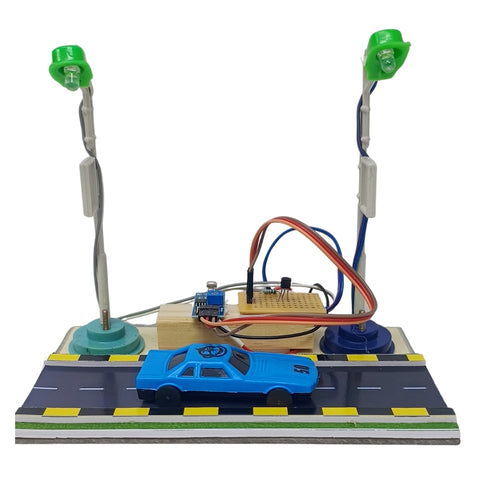 Smart Street Light school project  bu Go Science.A small electronic toy car setup on a road base with two green-lit pole lights, a blue toy car, and a central circuit board with wires, photographed on a plain white background.