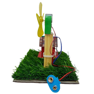 A small wind turbine model with a yellow plastic fan, green and silver blades, mounted on a wooden pole over artificial green grass, with red and blue wires leading to a blue battery connector on a white background. Windmill Generator (Windturbine) School Project go science classroom series image 6